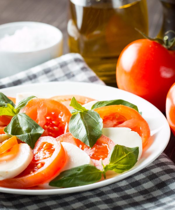 Photo of Caprese Salad with tomatoes, basil, mozzarella, olives and olive oil on wooden background. Italian traditional caprese salad ingredients. Mediterranean, organic and natural food concept.